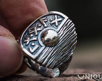 May include: A silver ring with a shield design featuring a sun and runes. The shield is textured and has a raised edge.