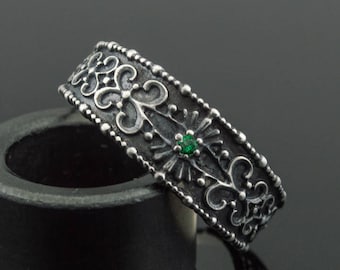 May include: A silver ring with an ornate design featuring a central green gemstone. The ring has a detailed pattern of swirls and dots. The ring is displayed on a black cylindrical object against a dark background.