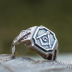 K&ouml;nnte beinhalten: Ein silberner Ring mit einem freimaurerischen Symbol, einem Zirkel und einem Winkelmesser, auf der Vorderseite. Der Ring ist mit komplizierten Designs verziert und liegt auf einem grauen Stein.