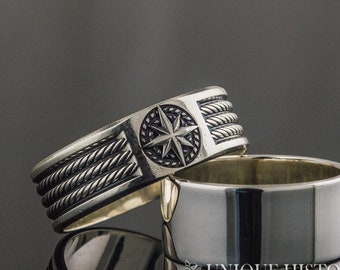 May include: A silver ring with a compass rose design and a rope-like band. The ring is on a gray surface.
