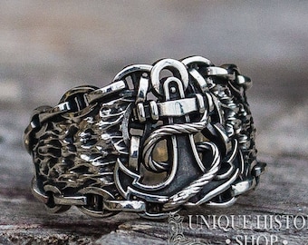 May include: A silver ring with a detailed anchor design. The anchor is surrounded by a textured band of linked chains.