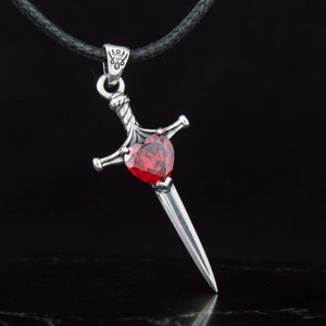 May include: A silver pendant necklace with a sword design. The sword has a red heart-shaped gemstone in the center.