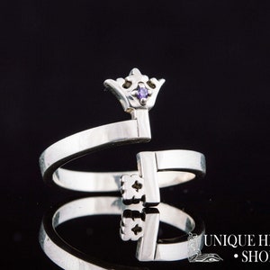 May include: A silver ring with a crown-shaped key design. The key has a small purple gemstone in the crown.