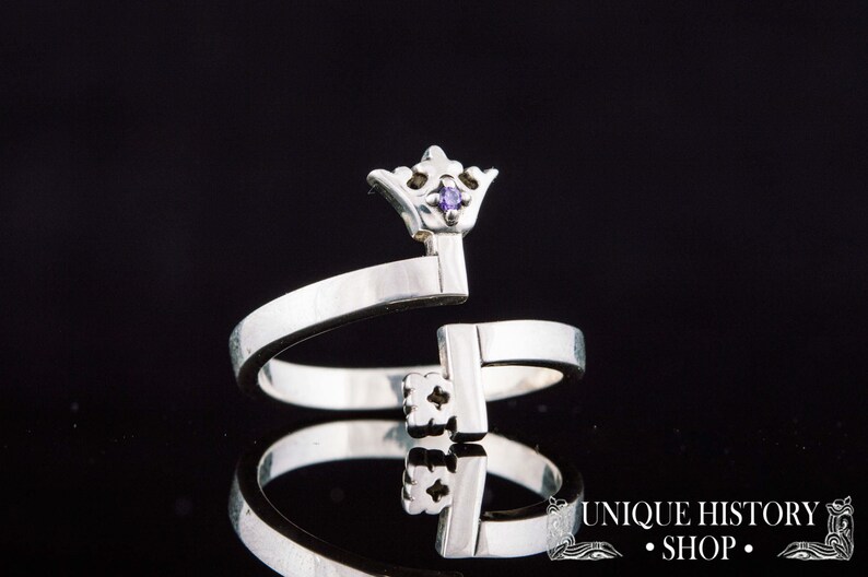 May include: A silver ring with a crown-shaped key design. The key has a small purple gemstone in the crown.