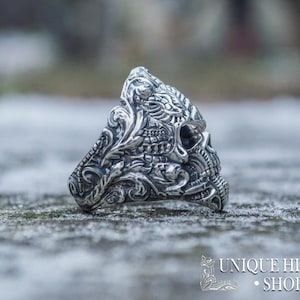Peut inclure: Bague en argent d&eacute;taill&eacute;e avec un motif de cr&acirc;ne et des motifs floraux et ornementaux complexes. La conception de la bague comprend un cr&acirc;ne avec des sculptures d&eacute;taill&eacute;es, sur un fond flou et enneig&eacute;. La bague est de Unique History Shop.