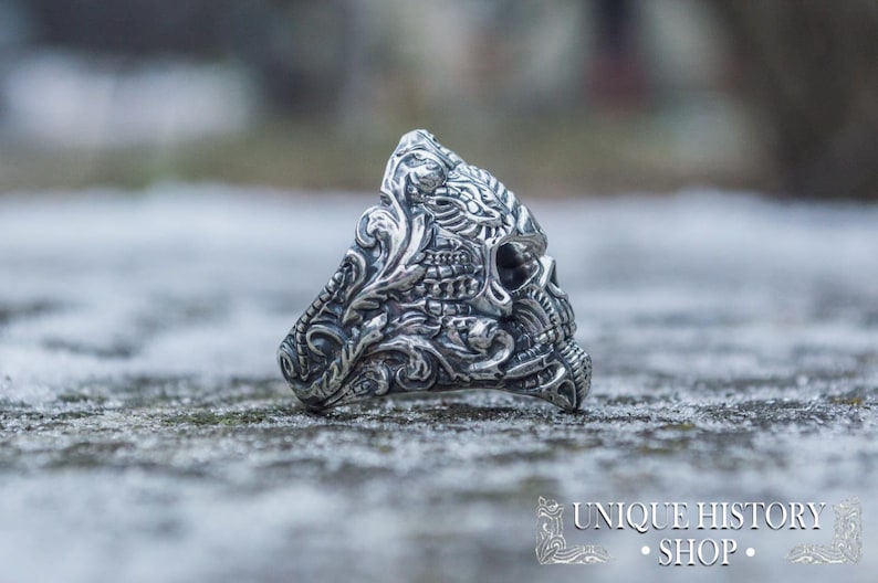 Peut inclure: Bague en argent d&eacute;taill&eacute;e avec un motif de cr&acirc;ne et des motifs floraux et ornementaux complexes. La conception de la bague comprend un cr&acirc;ne avec des sculptures d&eacute;taill&eacute;es, sur un fond flou et enneig&eacute;. La bague est de Unique History Shop.