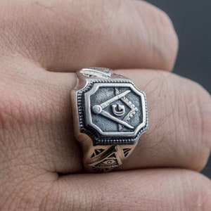 K&ouml;nnte beinhalten: Ein silberner Ring mit einem quadratischen Design, das einen Zirkel und ein Geodreieck zeigt, ein Symbol der Freimaurerei.