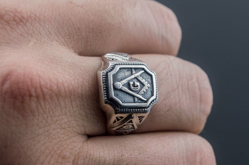 K&ouml;nnte beinhalten: Ein silberner Ring mit einem quadratischen Design, das einen Zirkel und ein Geodreieck zeigt, ein Symbol der Freimaurerei.