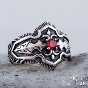 May include: A silver ring with an ornate design featuring a central red gemstone. The ring has a detailed, textured pattern with arrow-like shapes and floral accents. The ring is set against a neutral background.