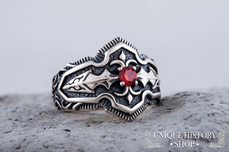 May include: A silver ring with an ornate design featuring a central red gemstone. The ring has a detailed, textured pattern with arrow-like shapes and floral accents. The ring is set against a neutral background.