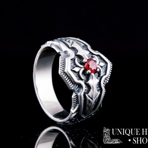 May include: A silver ring with an ornate design featuring a central red gemstone. The ring has a detailed, textured pattern and is set against a black background. The text "UNIQUE HISTORY SHOP" is visible in the lower right corner.