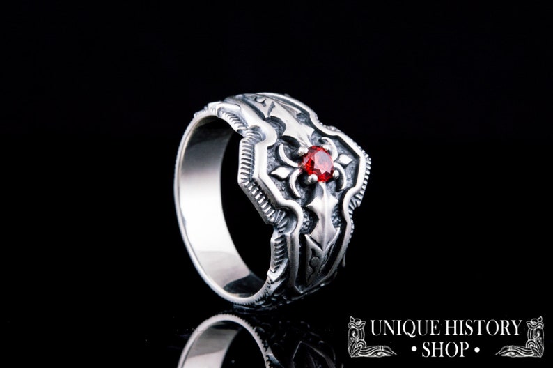 May include: A silver ring with an ornate design featuring a central red gemstone. The ring has a detailed, textured pattern and is set against a black background. The text "UNIQUE HISTORY SHOP" is visible in the lower right corner.