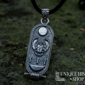 May include: Silver pendant featuring a scarab beetle with wings, a sun disc, and hieroglyphs. The pendant is on a black cord and is resting on a textured surface.