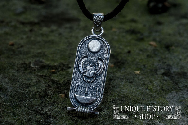 May include: Silver pendant featuring a scarab beetle with wings, a sun disc, and hieroglyphs. The pendant is on a black cord and is resting on a textured surface.