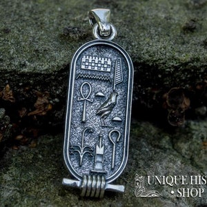 May include: Silver pendant with an Egyptian cartouche design. The cartouche features an ankh, a winged scarab beetle, and other hieroglyphs. The pendant is on a rocky surface.
