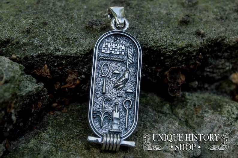 May include: Silver pendant with an Egyptian cartouche design. The cartouche features an ankh, a winged scarab beetle, and other hieroglyphs. The pendant is on a rocky surface.