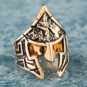Pode incluir: Um anel dourado com um design detalhado de um capacete espartano. O capacete &eacute; texturizado e tem um padr&atilde;o geom&eacute;trico ao redor das bordas.