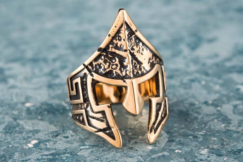 Pode incluir: Um anel dourado com um design detalhado de um capacete espartano. O capacete &eacute; texturizado e tem um padr&atilde;o geom&eacute;trico ao redor das bordas.