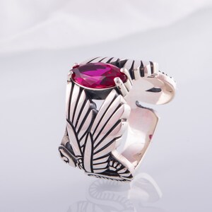 May include: A silver ring with a magenta marquise-cut gemstone. The ring has an openwork design with leaf patterns. The adjustable band is visible. The ring is set against a white background.
