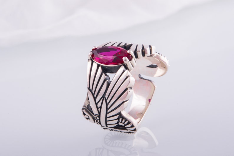 May include: A silver ring with a magenta marquise-cut gemstone. The ring has an openwork design with leaf patterns. The adjustable band is visible. The ring is set against a white background.