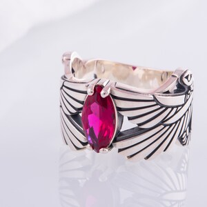 May include: A silver ring with a magenta gemstone in a marquise cut. The ring has a detailed wing design on the sides and decorative elements. The ring is displayed on a white surface.