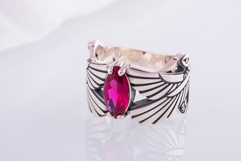 May include: A silver ring with a magenta gemstone in a marquise cut. The ring has a detailed wing design on the sides and decorative elements. The ring is displayed on a white surface.