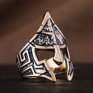 Pode incluir: Um anel de prata com um design detalhado de um capacete de guerreiro grego. O capacete &eacute; texturizado e tem um padr&atilde;o geom&eacute;trico ao redor das bordas.