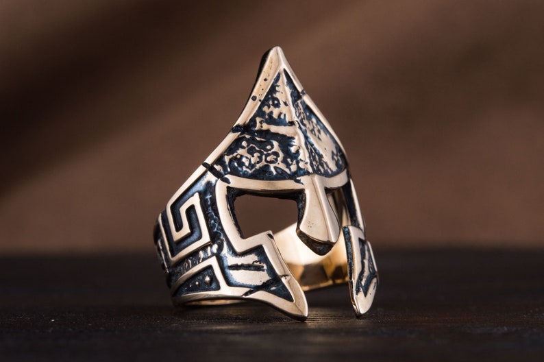 Pode incluir: Um anel de prata com um design detalhado de um capacete de guerreiro grego. O capacete &eacute; texturizado e tem um padr&atilde;o geom&eacute;trico ao redor das bordas.