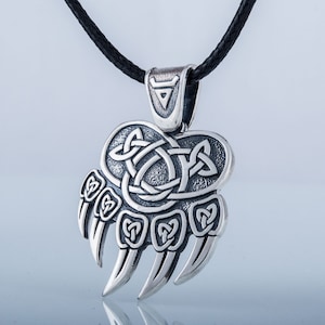 May include: Silver pendant necklace featuring a bear paw print with a Celtic knot design. The pendant is on a black cord.