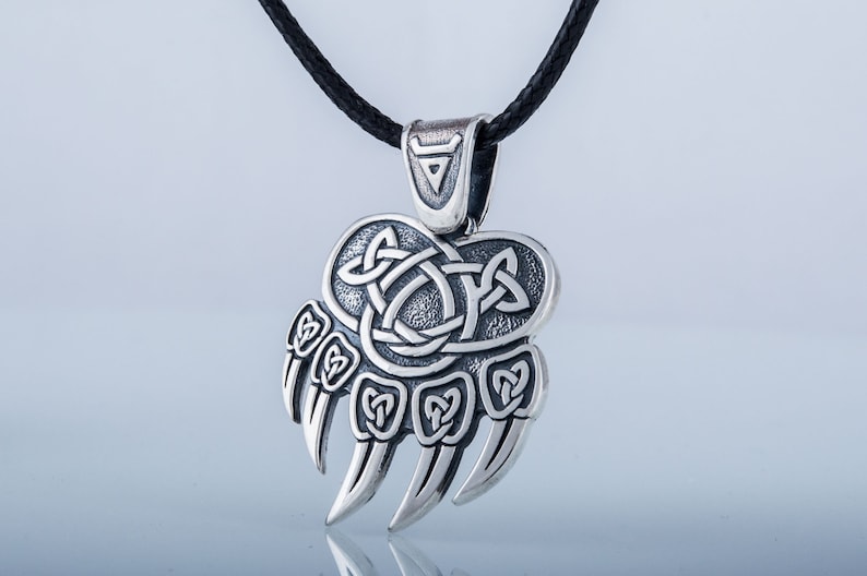 May include: Silver pendant necklace featuring a bear paw print with a Celtic knot design. The pendant is on a black cord.