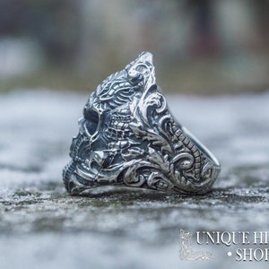 Peut inclure: Une bague argent&eacute;e avec un motif de cr&acirc;ne d&eacute;taill&eacute;. La bague pr&eacute;sente des gravures et des volutes complexes, avec le texte "UNIQUE HISTORY SHOP" en bas. La bague est pos&eacute;e sur un fond flou et textur&eacute;.