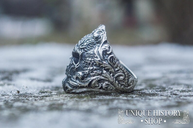 Peut inclure: Une bague argent&eacute;e avec un motif de cr&acirc;ne d&eacute;taill&eacute;. La bague pr&eacute;sente des gravures et des volutes complexes, avec le texte "UNIQUE HISTORY SHOP" en bas. La bague est pos&eacute;e sur un fond flou et textur&eacute;.