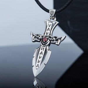 May include: A silver pendant in the shape of a sword with a red gemstone at the center. The sword has the words "Valhalla" inscribed on it.