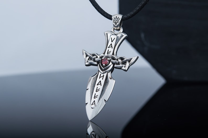 May include: A silver pendant in the shape of a sword with a red gemstone at the center. The sword has the words "Valhalla" inscribed on it.