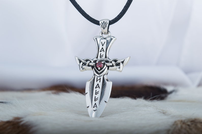 May include: Silver pendant necklace with a sword design and a red gemstone. The sword has the word "Valhalla" inscribed on it.