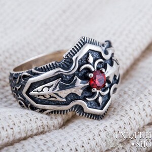 May include: A silver ring with an ornate design and a central red gemstone. The ring features detailed patterns and a textured finish. The ring is displayed on a white, textured surface. The ring is from the Unique History Shop.