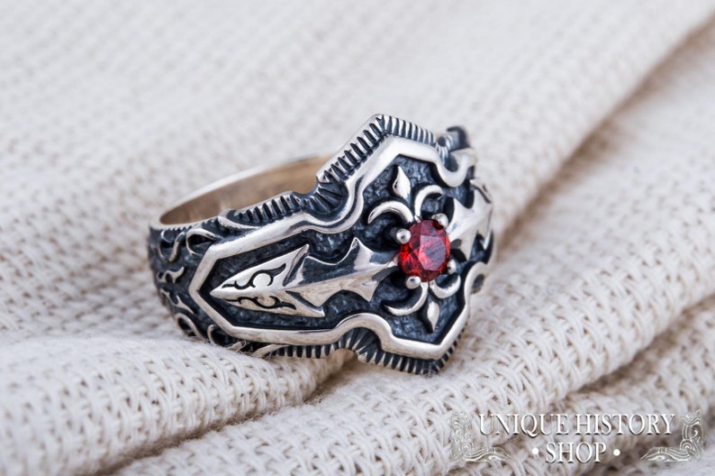 May include: A silver ring with an ornate design and a central red gemstone. The ring features detailed patterns and a textured finish. The ring is displayed on a white, textured surface. The ring is from the Unique History Shop.