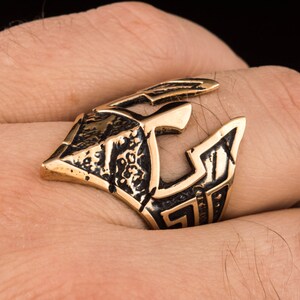 Pode incluir: Um anel dourado com um design detalhado de um capacete. O anel &eacute; usado no dedo de uma pessoa.