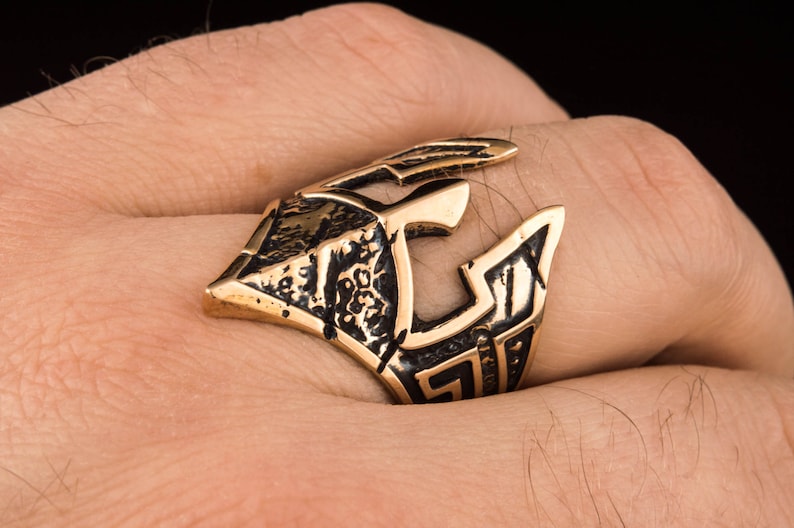 Pode incluir: Um anel dourado com um design detalhado de um capacete. O anel &eacute; usado no dedo de uma pessoa.