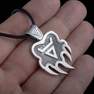 May include: Silver pendant necklace with a bear claw design and a triangle symbol inside. The pendant is on a black cord.