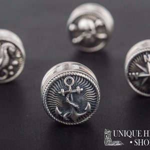 Puede incluir: Cuatro botones de metal plateados con diferentes dise&ntilde;os n&aacute;uticos. Los botones presentan una br&uacute;jula, un ancla, un caballito de mar y una calavera y huesos cruzados.