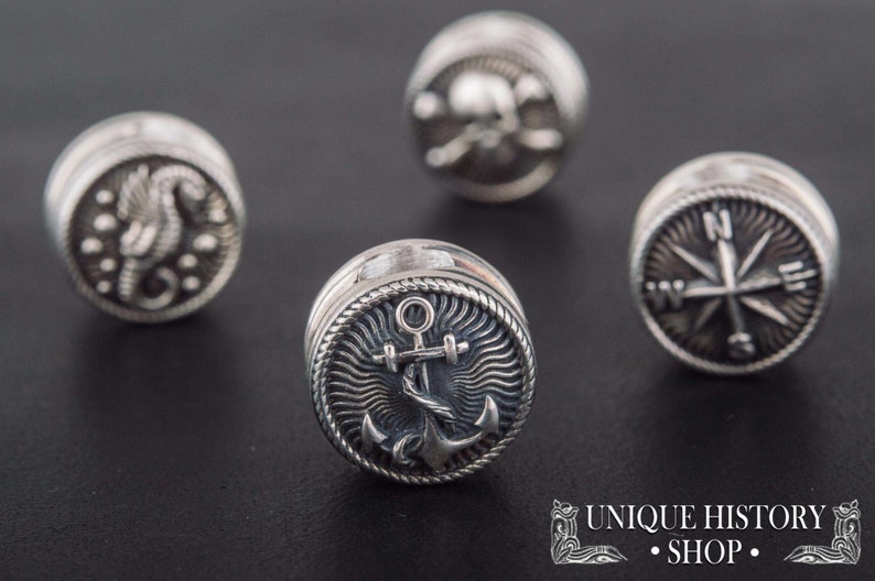 Puede incluir: Cuatro botones de metal plateados con diferentes dise&ntilde;os n&aacute;uticos. Los botones presentan una br&uacute;jula, un ancla, un caballito de mar y una calavera y huesos cruzados.