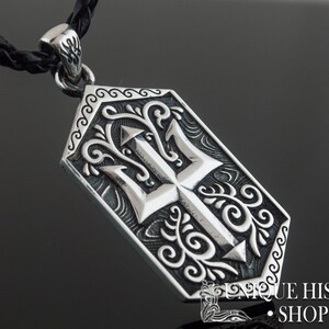 May include: Silver pendant necklace with a detailed trident design surrounded by ornate scrollwork. The pendant is attached to a black braided cord.