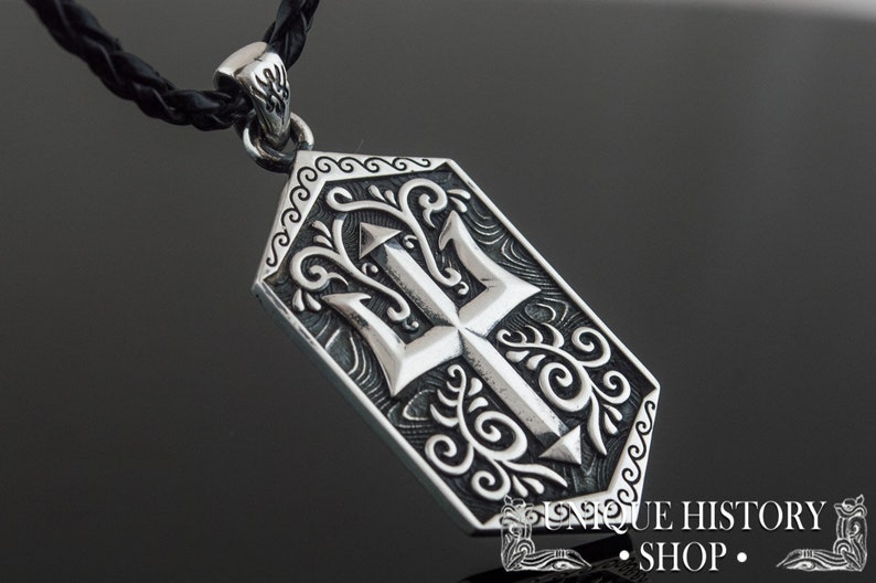 May include: Silver pendant necklace with a detailed trident design surrounded by ornate scrollwork. The pendant is attached to a black braided cord.