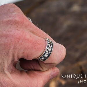 May include: A silver ring with a band of skulls. The ring is worn on a person's finger.
