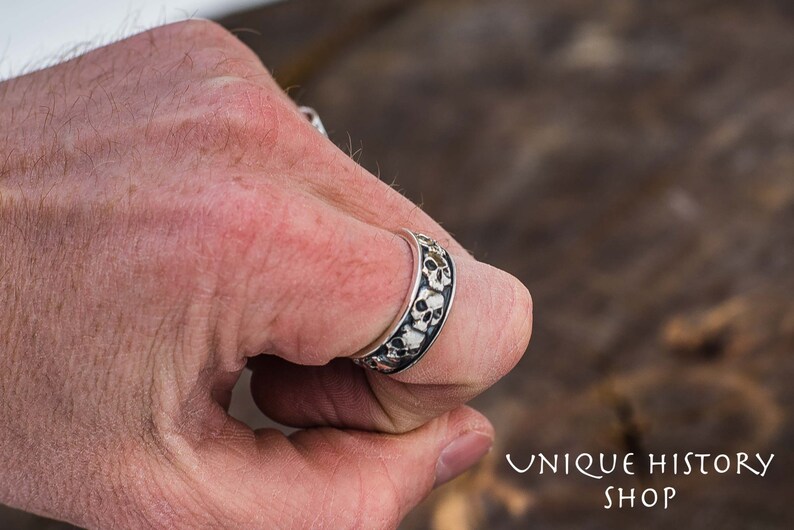 May include: A silver ring with a band of skulls. The ring is worn on a person's finger.