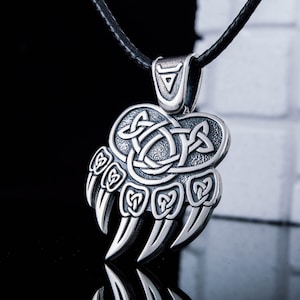 May include: A silver bear paw pendant necklace with a Celtic knot design. The pendant is attached to a black leather cord.