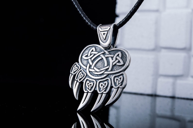 May include: A silver bear paw pendant necklace with a Celtic knot design. The pendant is attached to a black leather cord.