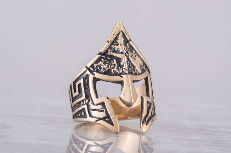 Pode incluir: Um anel dourado com um design detalhado de um capacete espartano. O capacete &eacute; texturizado e tem um padr&atilde;o de chave grega ao redor da banda.