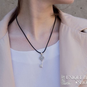 K&ouml;nnte beinhalten: Eine Halskette mit einem silbernen Schl&uuml;sselanh&auml;nger an einem schwarzen Band. Der Schl&uuml;ssel hat ein filigranes Design und ist ein beliebter Stil f&uuml;r Schmuck.